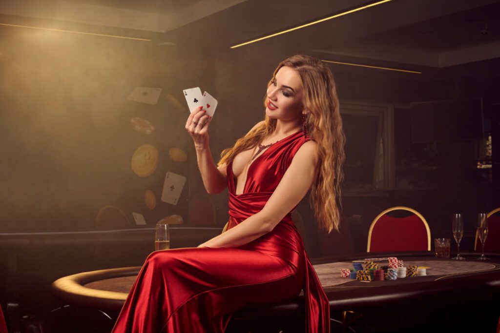 showy blond maiden long red satin dress with two aces her hand is posing sitting sideways poker table luxury casino passion cards chips alcohol win gambling it is female ent
